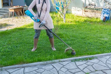 Fototapete Grün Woman gardener using electric string trimmer to cut grass in backyard. Lawn maintenance with garden equipment. Professional landscaping task and yard improvement work during spring  © Itana