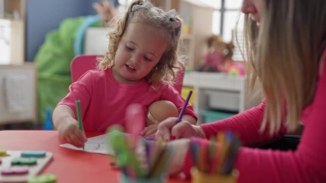 Little girl draws with crayon holding doll while woman guides hand at a table in a building; focused learning.