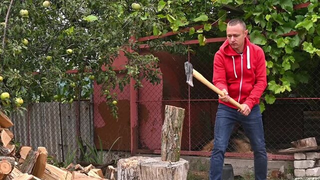 man splits a log with an axe, the log breaking in half after a strong second strike. He is wearing a red hoodie and jeans, natural greenery and wooden fence add to the rural atmosphere.
