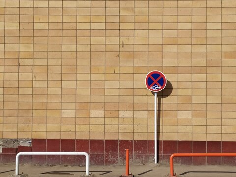 Beige tiled wall with red base featuring white pole with circular no parking sign and orange safety barriers in front creating strict urban regulation scene.