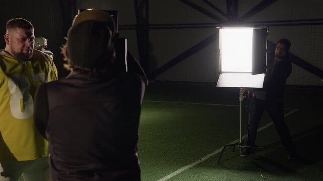 Medium backstage shot of Caucasian cameraman shooting video portrait or muscular American football player in yellow jersey at sports arena while directing gaffer to reposition light