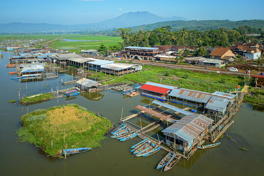 Aerial view of Spot Mancing Sumurup Rawapening with floating fishing huts, small blue boats, and a railway track near the lake in Semarang Regency, Central Java, Indonesia.