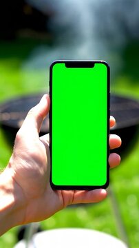 Hand Holding Smartphone with Green Screen Overlooking a Barbecue Grill with Smoke Rising on a Sunny Day