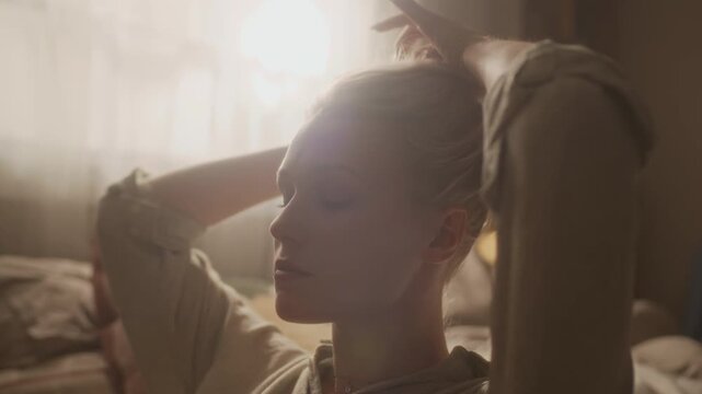 Handheld shot of young adult Caucasian girl tying hair into bun and leaving loose strand in cozy sunlit bedroom