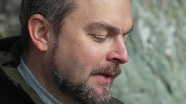 Man face close-up bearded male speaking outdoors against a rock wall in a pensive mood
