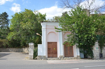 Obraz premium A small pink building with classical white arches and wooden doors located along an asphalt road. Lush green trees and a blue sky with clouds create a peaceful urban scene in summer day