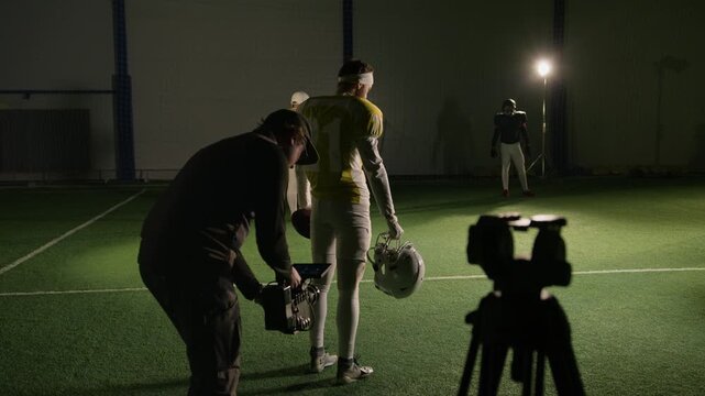 Full rear shot of cameraman filming American Football player in jersey, carrying helmet, on ground level walking across training arena towards team for practice, while shooting movie or commercial