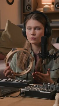 Vertical of focused young adult female musician recording tambourine shakes, shaping audio into samples while editing track in DAW on laptop in home studio