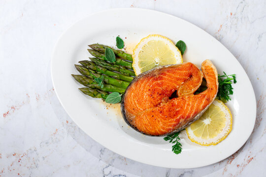 Salmon fillet with asparagus and lemon slices served on a white plate, light marble table, sunlight, home, no people,