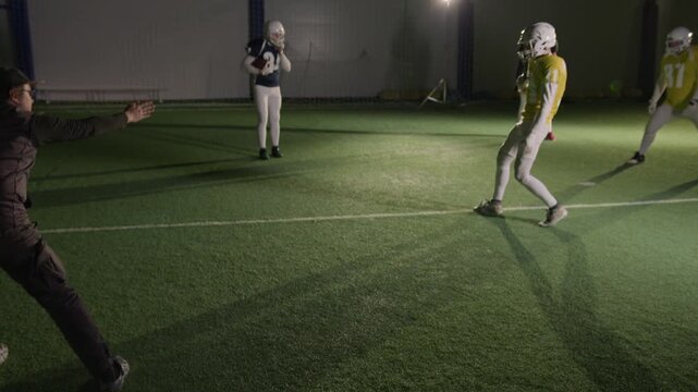 Full shot of Caucasian male cinematographer kneeling on turf to shoot American football team preparing to train at stadium, walking over to give instructions, while player tying shoelaces on cleats