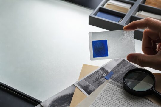 Hand holding an art or color sample card over a desk with documents and magnifying glass in a research workspace