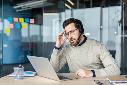 Man in headset at office desk holding his head in pain, stressed and fatigued during a tense customer support call or online meeting, struggling with workload and deadlines