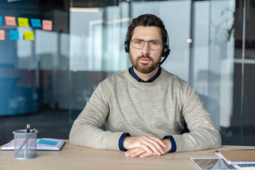 Fototapeta premium Male customer service representative wearing a headset and spectacles, looking directly at the camera while working in a modern office, representing professionalism and communication