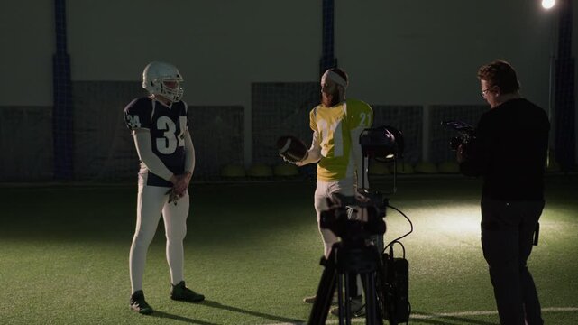 Full backstage shot of two Caucasian male American football players in team jerseys and helmets standing on pitch, talking and bouncing ball, videographer filming on handheld camera for commercial