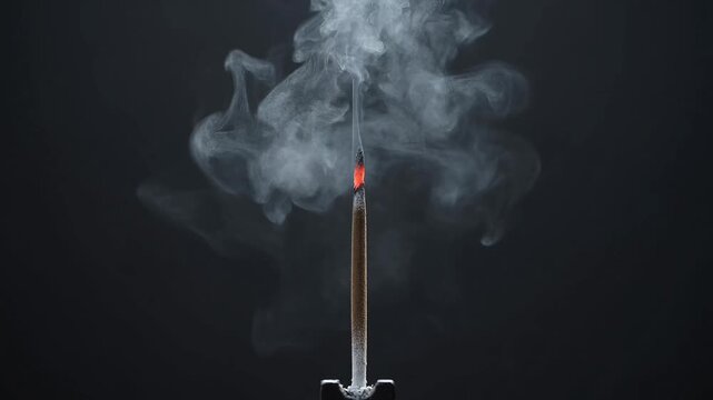 Single aromatic incense stick burning with graceful white smoke trails against a dark background