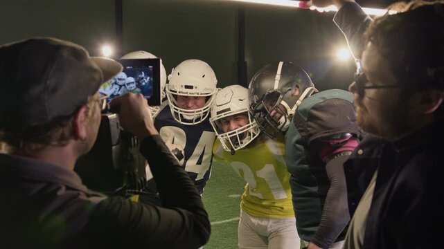 Medium arc backstage shot of TV production crew cameraman filming American football team players hugging after successful match, wearing protective helmets, gaffer holding light stick adobe heads