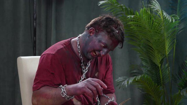An adult man in a zombie costume with bloody makeup and chain swipes a cardboard piece like a smartphone creating a humorous Halloween scene of horror and digital addiction.