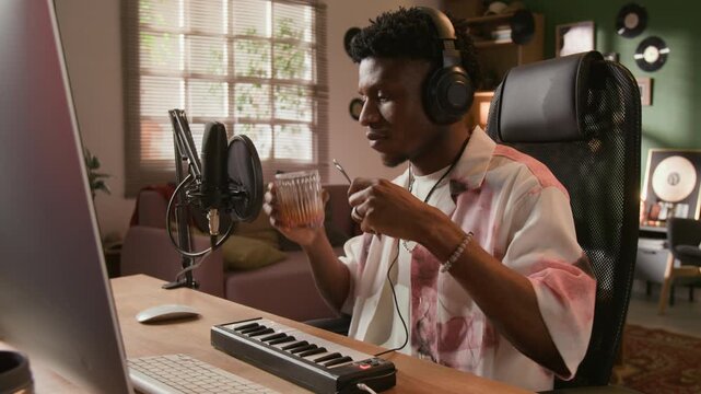 Medium shot of young adult African American music producer with headphones on recording metallic glass clinks in home based studio while creating experimental sound design