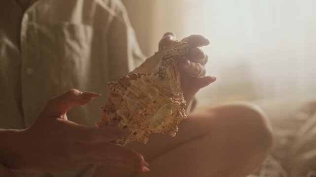 Cropped shot of unrecognizable woman holding and examining big seashell while sitting on bed under warm morning light