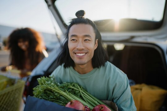 Young man carrying grocery bag smiling at open car trunk lot