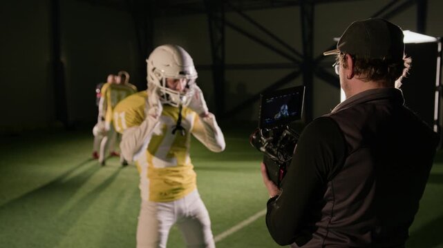 Medium rear behind scenes shot of busy male cinematographer giving directions to American football athlete, wearing body armor, team jersey, while filming sportsman putting on helmet for team practice
