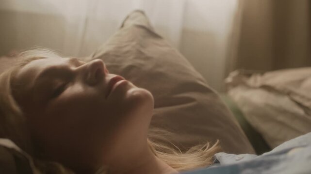 Close up portrait of gen Z Caucasian blonde woman untangling wired earphones and looking at camera while lying in bed under warm morning sunlight