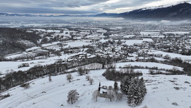 Aerial view of the snow-covered village of Vesancy and the historic Chapelle de Riantmont surrounded by winter fields and mountains in Vesancy, Auvergne-Rhone-Alpes, France.