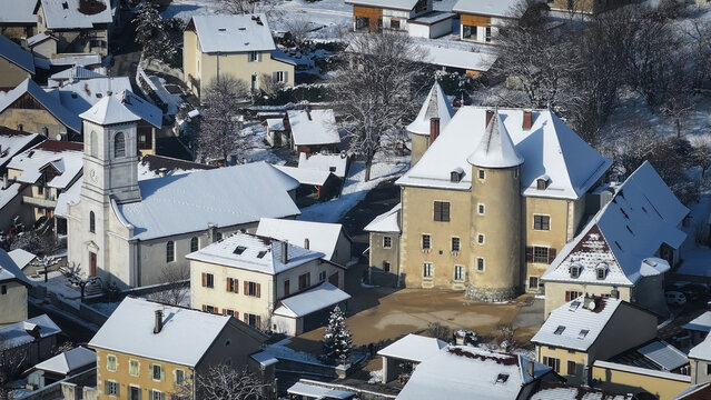Aerial view of the historic Vesancy castle and church surrounded by snow-covered rooftops and winter trees in Vesancy, Auvergne-Rhone-Alpes, France.