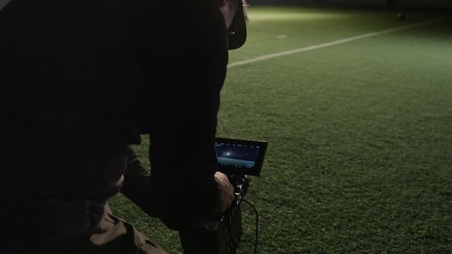 Medium full shot of skilled Caucasian male videographer kneeling on grass at sports arena, instructing gridiron football team before filming player running and falling during practice game for TV show