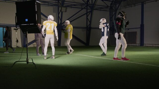 Wide backstage shot of Caucasian male videographer with camera shooting repeat takes of gridiron football players running with ball and tackling rival on sports field for commercial or TV show