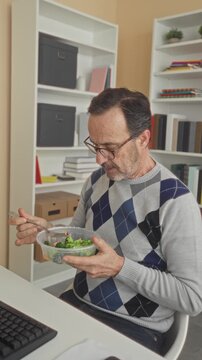 Middleaged hispanic man eating salad, holding plastic bowl and fork while seated at desk wearing argyle sweater in building; mindful healthy calm.