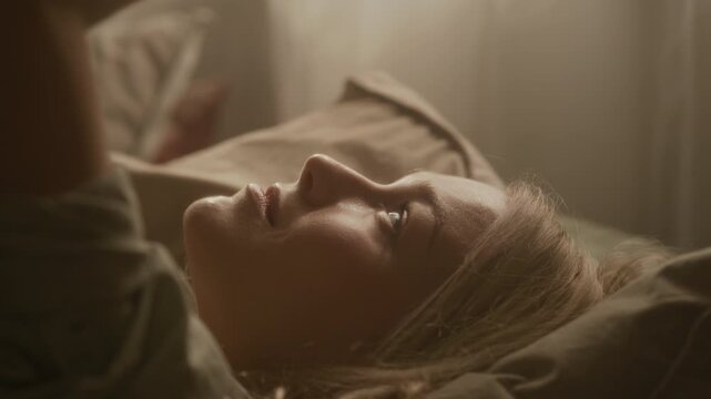 Close up shot of dreamy Caucasian gen Z woman lingering in bed after waking up in warm golden sunlight at home