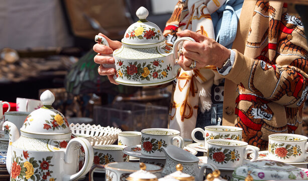 Woman holding decorative porcelain teapot while examining vintage dishware at a flea market, surrounded by various antique items and collectibles on display