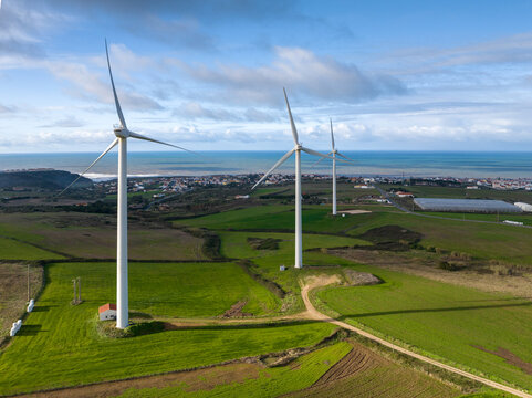 Aerial view of three wind turbines standing in green fields overlooking a coastal town and the Atlantic Ocean under a cloudy sky Ribamar, Portugal.