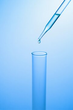 Close up of a glass pipette dropping clear liquid into a laboratory test tube on a blue background