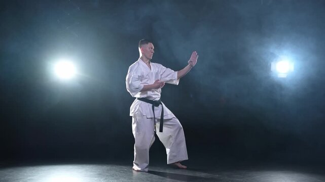 Strong man fighter in white kimono for karate martial arts with black belt preparing for fight training in black background. Man training moves punches kicks practicing shadow fight ready for sparring