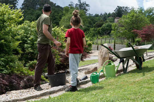 A man and a child stand together in a scenic backyard garden next to landscaping tools, working on an outdoor family project.