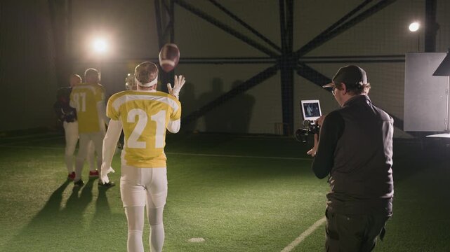 Full rear backstage shot of Caucasian cameraman following gridiron football player in yellow jersey bouncing ball, walking towards team mates across arena, then cutting and returning for another take