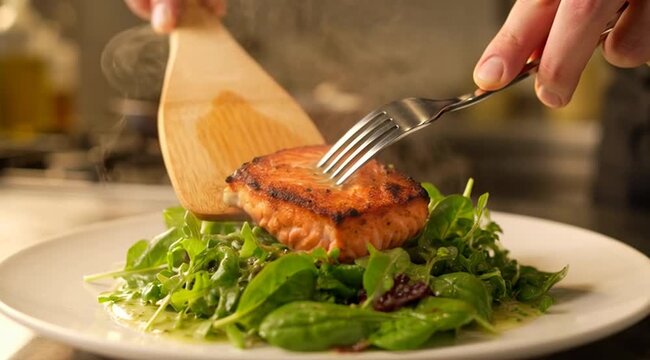 Chef places a seared salmon fillet onto a fresh green salad with a wooden spatula, showcasing gourmet cooking, culinary artistry, healthy eating, and meal plating