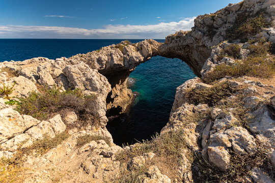 Breathtaking natural rock arch formation spanning the clear turquoise sea against a rugged Mediterranean coastline under a vibrant blue sky.