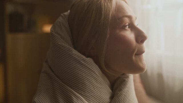 Close up shot of dreamy young girl sitting on bed and wrapping herself in soft blanket under warm morning light