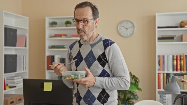 Man wearing argyle sweater holds salad container and fork while eating at office desk inside building, looking at computer screen; calm focus.