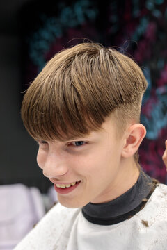 close up portrait of smiling teenage boy with bowl cut fade hairstyle looking slightly down at camera with blue eyes in barbershop setting