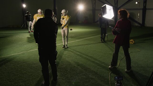 Wide shot of production crew shooting American football team training at stadium, setting up kickoff scene, directing players to assume positions on field, while gaffer is installing lights