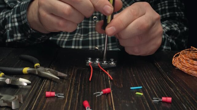 Hands of a male technician using a screwdriver to connect wires to a terminal block on a wooden workbench with tools and connectors nearby