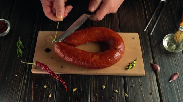 The hands of a chef cutting a sausage string on a wooden cutting board are surrounded by spices, a knife and kitchen utensils, creating a vibrant culinary scene