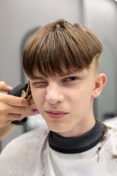 close up portrait of teenage boy with bowl cut winking one eye while barber applies trimmer near temple in grey modern barbershop