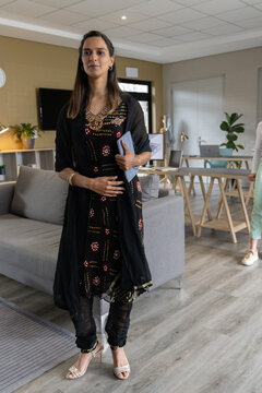 Coworkers with Indian woman in black kurta standing near grey sofa in office, holding blue tablet