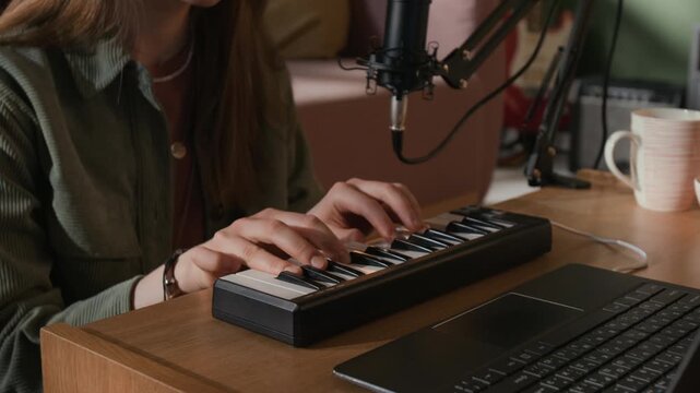 Close up on hands of young adult female composer playing MIDI keyboard at production setup with laptop while recording electronic music in home studio