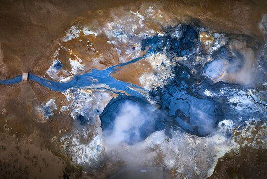 Aerial view of a geothermal field featuring vibrant blue mud pools, rising steam vents, and mineral-rich orange earth across the volcanic landscape of Iceland.